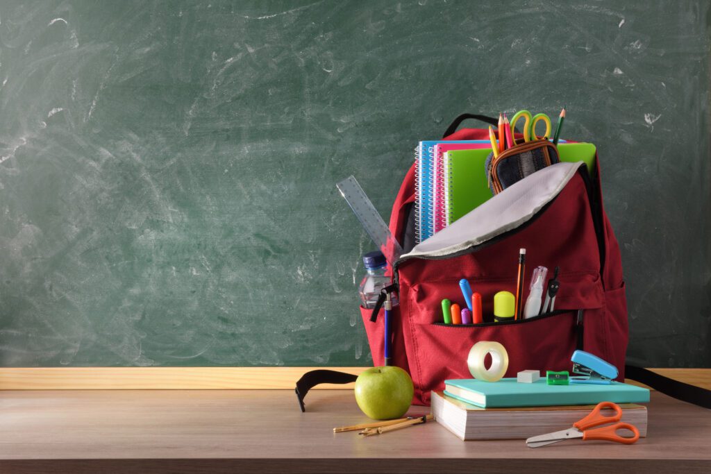 Backpack full of school supplies on wood table and green blackboard | Image credit: Davizro Photography - stock.adobe.com